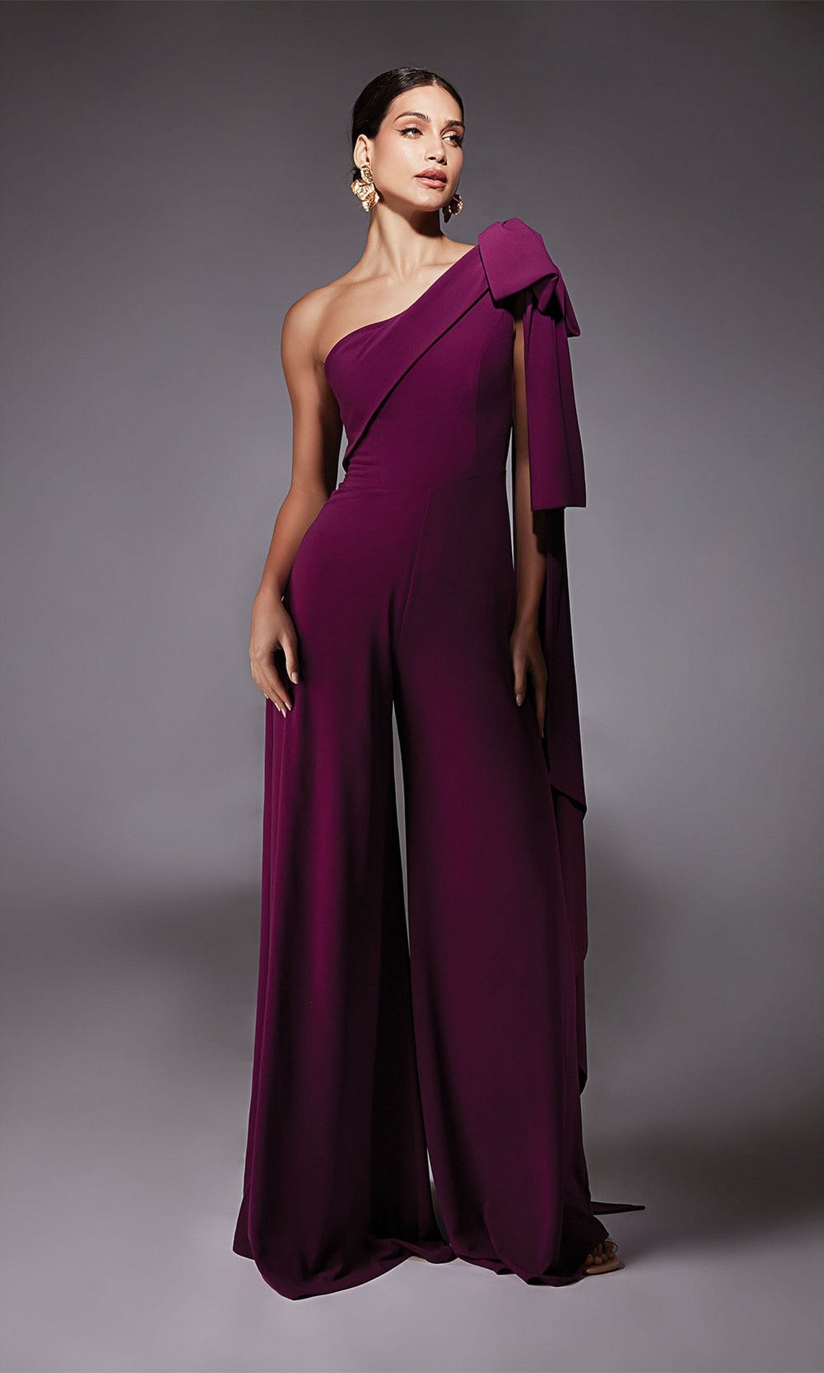 A woman wears the Alyce 70065, a long formal purple one-shoulder jumpsuit with a large bow detail and wide-leg pants, paired with gold statement earrings. She stands against a grey background—a chic alternative to a cocktail dress.